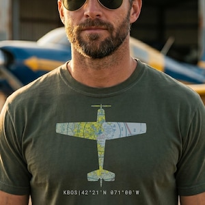 May include: An olive green short-sleeve t-shirt featuring a graphic of an airplane silhouette filled with a colorful aeronautical map pattern. Below the airplane graphic, the text reads KBOS | 42°21'N 071°00'W in a clean white sans-serif font.