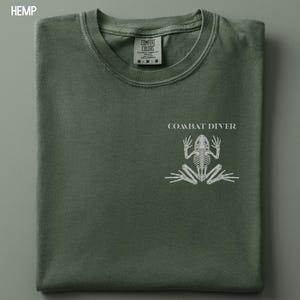 May include: A hemp-colored crew neck t-shirt featuring a white graphic of a frog skeleton on the chest. Above the skeleton, the text reads COMBAT DIVER. The interior neck label displays the brand name Comfort Colors and the size M.