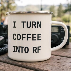 May include: A white enamel mug with a silver-coloured rim and a handle on the right side. The front of the mug features the black printed text I TURN COFFEE INTO RF in a bold, sans-serif font. The mug has a smooth, glossy finish throughout.