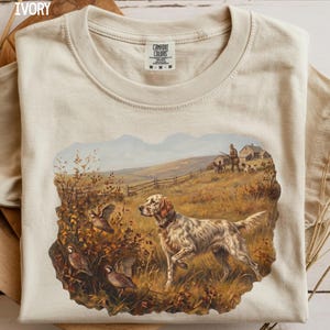 May include: An ivory t-shirt featuring a graphic print of a hunting scene. The illustration depicts a white and brown spotted hunting dog in a field with several birds, a person with a rifle, and a small building in the background. The shirt has a crew neckline and a Comfort Colors brand label at the inner neck.