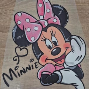 Könnte beinhalten: Ein farbenfrohes Bild von Minnie Mouse mit einer rosa Schleife mit weißen Punkten, einem rosa Kleid und weißen Handschuhen. Das Wort "Minnie" ist kursiv mit einem Herzsymbol geschrieben.