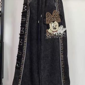 Könnte beinhalten: Schwarze Acid-Wash-Jogginghose mit elastischem Kordelzugbund. An der Seite befindet sich ein vertikales schwarzes Paneel mit dem Wort MINNIE in weißen Buchstaben. Ein Minnie-Maus-Motiv mit Leoparden-Schleife und dem Wort FASHION ist vorne.
