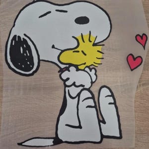 Peut inclure: Graphique de dessin animé représentant Snoopy embrassant Woodstock, avec deux cœurs rouges sur fond transparent. Snoopy est blanc avec des détails noirs. Woodstock est jaune. L'image est sur une surface brun clair.