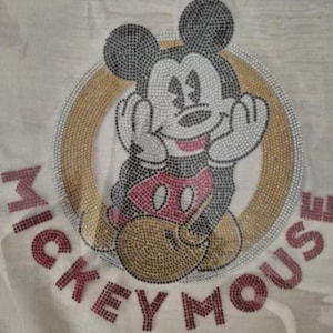 Könnte beinhalten: Nahaufnahme eines weißen Stoffes mit einem Mickey Mouse-Design. Das Design zeigt Mickey Mouse in sitzender Pose, umgeben von einem goldenen und silbernen Kreis. Die Worte "MICKEY MOUSE" sind in rosa Buchstaben darunter geschrieben.