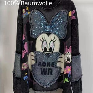 Könnte beinhalten: Ein schwarzes, säuregewaschenes Langarm-Sweatshirt mit einer großen, mit Pailletten besetzten Minnie-Maus-Grafik und einem herzförmigen Aufnäher mit dem Text AONE WR. Das Kleidungsstück enthält zusätzliche bunte Charakterdrucke, Blumenmotive und abgenutzte Ärmeleinsätze im Denim-Stil.
