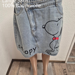 Könnte beinhalten: Ein hellblauer Jeansrock mit asymmetrischem Überlappungsbund, ausgefranstem Saum und einem Snoopy-Grafikdruck mit rotem Halsband. Der Text lautet: Einheitsgröße 44, Hosenbund 44cm, Länge 38cm, 100% Baumwolle. Der Rock hat seitliche Taschen.