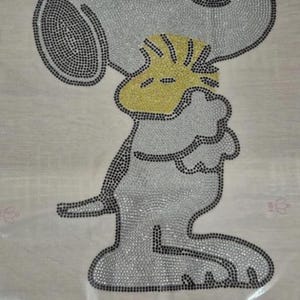 Peut inclure: Un motif Snoopy composé de strass argentés et noirs sur un fond clair. Le mot "SNOOPY" est épelé en strass noirs en bas de l'image. Le motif représente Snoopy en train d'embrasser Woodstock.