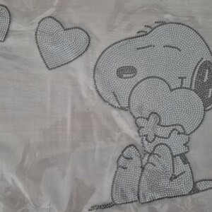 Könnte beinhalten: Ein hellgraues Snoopy-Design mit drei Herzen. Snoopy hält ein Herz und hat die Augen geschlossen. Das Design besteht aus kleinen, runden, grauen Punkten. Der Hintergrund ist ein hellgrauer Stoff.