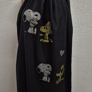 Könnte beinhalten: Ein schwarzer Schal mit Strassverzierungen von Snoopy und Woodstock. Das Design zeigt zwei Paare der Figuren aus silbernen und gelben Kristallen sowie zwei kleine herzförmige Strassakzente in Silber im oberen und mittleren Bereich des Schals.