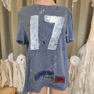May include: A blue t-shirt with the number "17" in white and blue stripes. The shirt has white and black paint splatters and patches of fabric in blue, green, and red near the hem. The shirt is displayed on a mannequin.