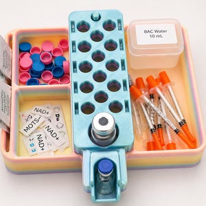 May include: A multi-compartment tray in pastel colors, holding various medical supplies. Includes syringes with orange caps, small vials, pink and blue caps, and packets labeled "Alcohol Prep Pads". A container reads "BAC Water 10 mL".