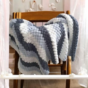 May include: A chunky knit blanket in shades of white, light blue, and dark gray is draped over a wooden chair. The blanket features a woven pattern and a soft, plush texture, creating a cozy and inviting aesthetic. The blanket is a home decor item.