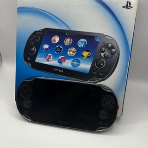 Può includere: Una console di gioco portatile Sony PlayStation Vita nera con la sua scatola. La console ha uno schermo che mostra un'interfaccia di gioco. La scatola presenta il logo Sony e un'immagine della console.