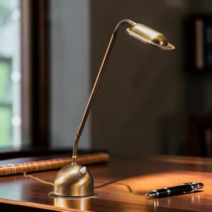 May include: A vintage-style desk lamp with a brass finish. The lamp features a rounded base, a slender, adjustable arm, and a rectangular light head. A black pen rests on the wooden desk.