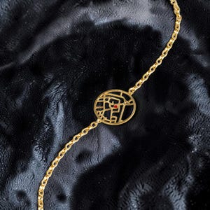 May include: A gold-toned chain bracelet featuring a circular pendant with an intricate, openwork street map design. The center of the map includes a small, singular red dot accent, and the chain consists of uniform, rounded metal links.