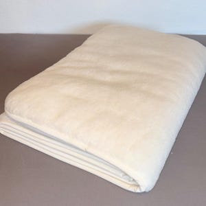 Pode incluir: Uma manta retangular espessa de cor branco-sujo com uma textura macia, felpuda e semelhante a lã polar. A manta está cuidadosamente dobrada numa pilha, exibindo a sua superfície de pelúcia e as bordas limpas e bem acabadas.