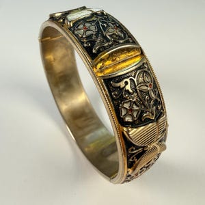 Puede incluir: Una pulsera rígida con bisagra, acabado en oro metálico e incrustaciones de esmalte negro. La superficie está decorada con motivos florales en relieve, patrones de hojas texturizadas y pequeños acentos rojos. El diseño incluye un patrón floral.