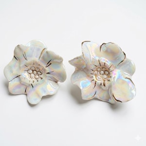 Könnte beinhalten: Ein Paar blumenförmige Keramikohrringe mit irisierendem, perlmuttartigem Finish, goldfarbenen Rändern und goldfarbenen Staubgefäßen in der Mitte. Die Blütenblätter haben eine wellige, organische Form mit einer glänzenden Oberfläche.