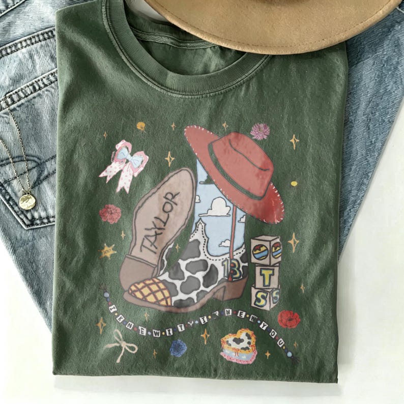 May include: An olive green t-shirt featuring a graphic print of a cowboy boot with a cow print pattern, a red hat, a pink bow, and blocks spelling TS. The text TAYLOR and 13 are visible on the boot, with the phrase I KNEW YOU WERE TROUBLE printed below.