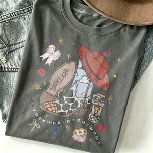 May include: A charcoal gray t-shirt featuring a graphic print of a cow-print cowboy boot, a red cowboy hat, a pink bow, and blocks with the text TS. The boot includes the name Taylor and the number 13, surrounded by stars and beads.
