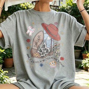 May include: A heather gray t-shirt featuring a graphic print of a cowboy boot with a cow print pattern, a red cowboy hat, a blue sky with clouds, a stack of blocks with the letters T, S, and the number 13, a pink bow, and text reading TAYLOR.