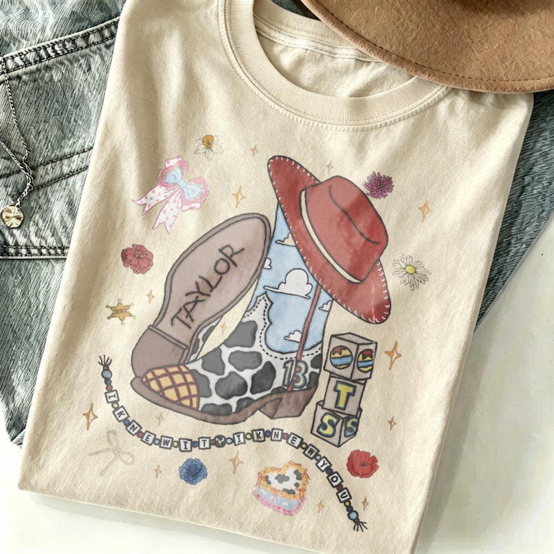 May include: A cream-colored t-shirt featuring a graphic print of a cow-print cowboy boot with a blue sky pattern, a red cowboy hat, a stack of blocks spelling BTS, a pink bow, flowers, and a beaded string with the text I KNEW IT Y I KNEW IT Y D E.