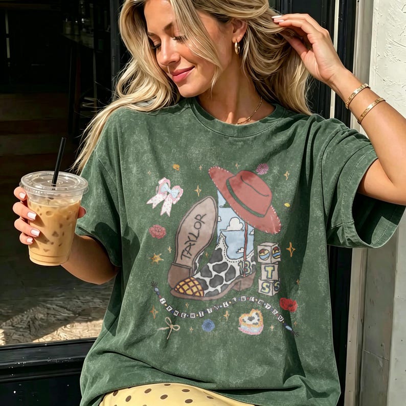 May include: A forest green mineral wash t-shirt featuring a graphic print of a cow print cowboy boot with the name Taylor, a red cowboy hat, a light blue bow, and stacked blocks with the letters T, S, and the number 13, surrounded by small stars.