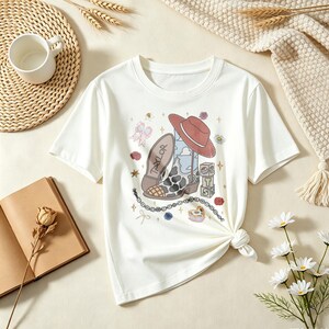 May include: A white short-sleeve t-shirt featuring a graphic print of a cowboy boot with a cow print pattern, a red hat, a stack of blocks with the letters T, S, and 13, a chain, and small floral and bow accents on the front center.