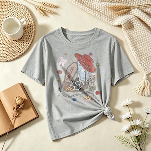 May include: A heather gray short-sleeve t-shirt featuring a graphic print of a cowboy boot with a cow-print pattern, a red hat, and blocks labeled with the letters T, S, and the number 13. The word Taylor is printed on the boot sole.