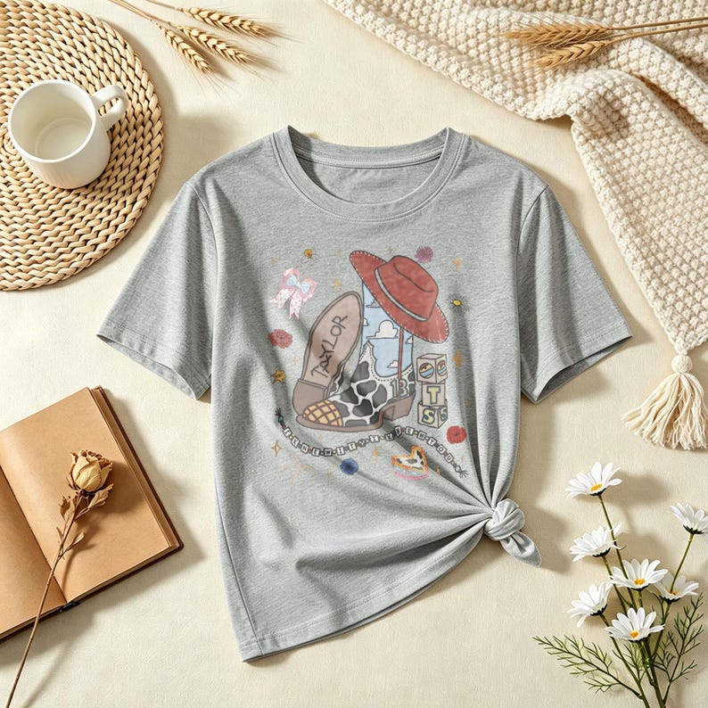 May include: A heather gray short-sleeve t-shirt featuring a graphic print of a cowboy boot with a cow-print pattern, a red hat, and blocks labeled with the letters T, S, and the number 13. The word Taylor is printed on the boot sole.