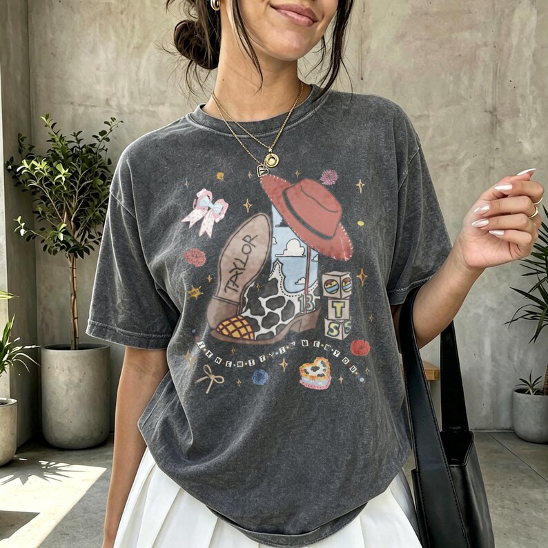 May include: A charcoal gray short-sleeve t-shirt featuring a graphic print of a cowboy boot with a cow print pattern, a red hat, and blocks spelling T-S-T. The boot has the name Taylor printed on the sole, surrounded by small stars and bows.