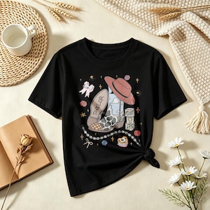 May include: A black short-sleeve t-shirt featuring a graphic print of a cowboy boot with a cow print pattern, a pink hat, a stack of blocks with the letters T, S, and a number 13, a bow, and the name Taylor written on the boot sole.