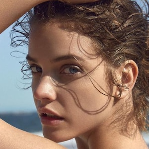Puede incluir: Retrato en primer plano de una persona con piel clara, ojos azules y cabello castaño mojado y despeinado. El brazo de la persona está levantado, y el fondo muestra un cielo azul y un mar borrosos. La iluminación crea sombras en la cara.