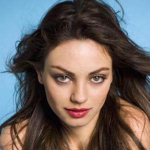 May include: A close-up portrait of a person with long, dark brown hair against a light blue background. The person has green eyes, and red lipstick. The hair is styled with some strands blowing in the wind.