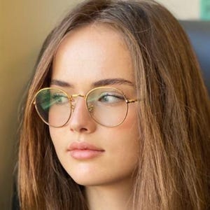 May include: A woman wearing gold-rimmed, round eyeglasses. The glasses have a thin frame and clear lenses. The woman has long, light brown hair and is looking to the side. The background is blurred, with hints of yellow and black.