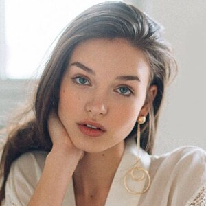 May include: A portrait of a person with long brown hair and blue eyes, wearing a white robe and gold earrings. The earrings have a round stud and a long chain with two connected circles.