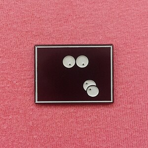 May include: A rectangular enamel pin with a dark burgundy background and a white border. The pin features two white eyes at the top and three white circles at the bottom, creating a minimalist design.