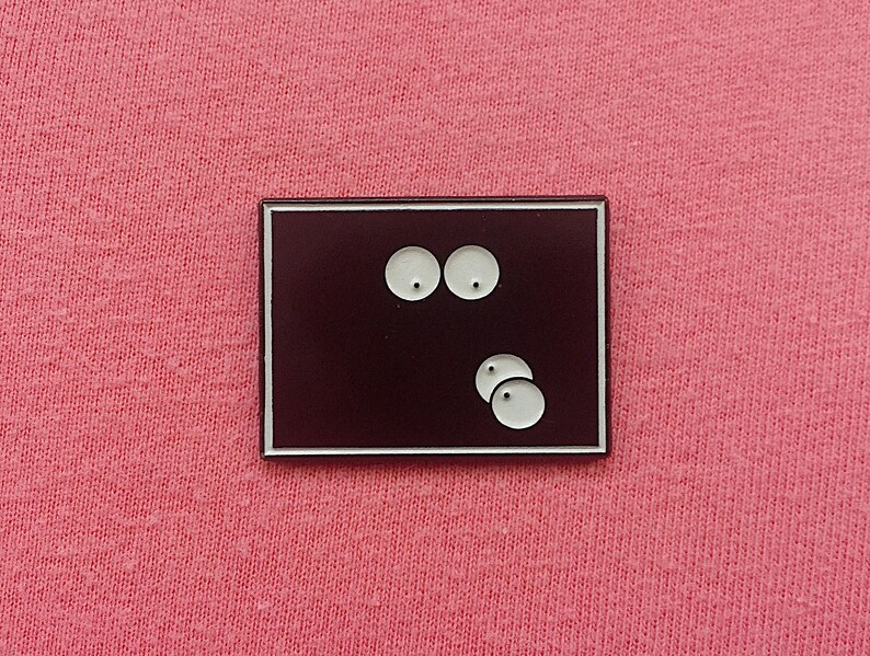May include: A rectangular enamel pin with a dark burgundy background and a white border. The pin features two white eyes at the top and three white circles at the bottom, creating a minimalist design.