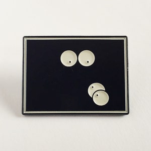 May include: A rectangular black enamel pin with a silver border. The pin features a minimalist design of two large, round, off-white eyes above three smaller, similar circles. The design is simple and graphic.