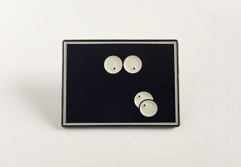 May include: A rectangular black enamel pin with a silver border. The pin features a minimalist design of two large, round, off-white eyes above three smaller, similar circles. The design is simple and graphic.