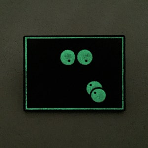May include: A black rectangular patch with a glowing green border and cartoon eyes. The eyes are two large circles above two smaller circles, all glowing green. The patch is on a gray surface.