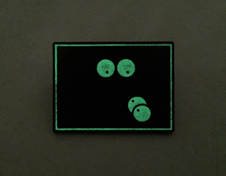 May include: A black rectangular patch with a glowing green border and cartoon eyes. The eyes are two large circles above two smaller circles, all glowing green. The patch is on a gray surface.