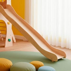 以下が含まれることがあります： 明るい茶色の木製滑り台。滑り台は、左側にクライミングウォールが見える、より大きな遊び構造の一部です。滑り台は、カラフルなフロアクッションが付いた緑色のマットの上にあります。