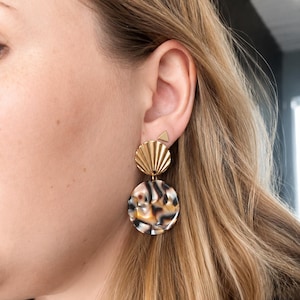 Peut inclure: Une boucle d'oreille en métal doré composée d'un petit clou triangulaire, d'une pièce supérieure texturée en forme d'éventail et d'un pendentif circulaire pendant avec un motif marbré dans des tons marron, noir, blanc et ambre.