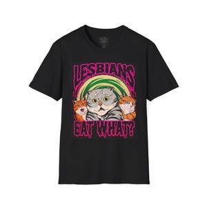 Puede incluir: Una camiseta negra de manga corta con un estampado gráfico de tres gatos con los ojos muy abiertos. El texto "LESBIANS EAT WHAT?" aparece en una fuente rosa que gotea sobre y debajo de los gatos, con un fondo circular verde y amarillo.