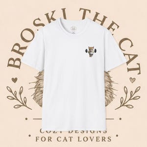 Könnte beinhalten: Ein weißes kurzärmeliges T-Shirt mit einer kleinen Grafik auf der linken Brust, die ein orangefarbenes Kätzchen beim Heben einer schwarzen Hantel zeigt. Im Hintergrund steht der Text BROSKI THE CAT und COZY DESIGNS FOR CAT LOVERS in Braun.