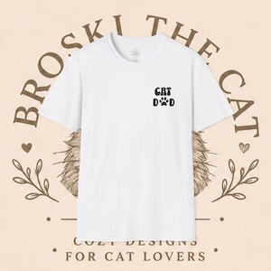 Puede incluir: Una camiseta blanca de manga corta con un gráfico negro en el pecho izquierdo que dice CAT DAD, con una huella de pata reemplazando la letra A. La camiseta tiene cuello redondo y un corte estándar sobre un fondo beige claro.