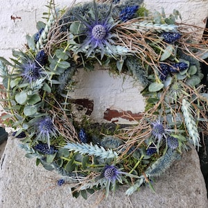 May include: A circular wreath composed of various dried plants and flowers. The wreath features a mix of green eucalyptus leaves, blue thistle flowers, and dried wheat. The arrangement is accented with twigs and moss, creating a natural, rustic aesthetic.
