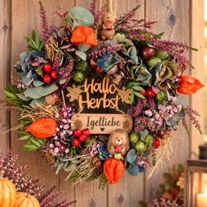 Può includere: Una ghirlanda autunnale circolare con erica viola essiccata, ortensie bluastre, lanterne di physalis arancioni e fogliame verde. Due piccole statuette di riccio sono inserite nella composizione. Un cartello in legno riporta il testo Hallo Herbst Igelliebe.