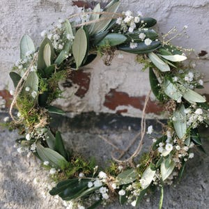 Peut inclure: Une couronne circulaire composée de branches d'olivier, de petites fleurs blanches et de mousse verte. La couronne est attachée avec de la ficelle et posée contre un mur de briques blanches patinées. Esthétique rustique et organique.